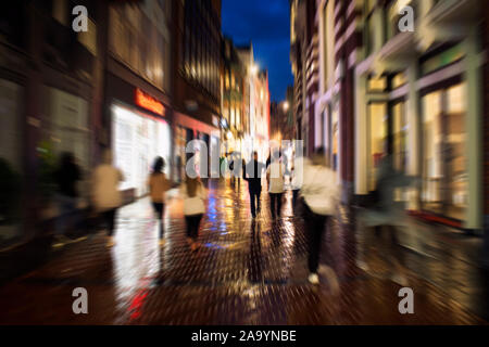 Blurry motion Image der jungen Menschen zu Fuß auf der Kalverstraat Straße, einer der wichtigsten Einkaufsstraßen in Amsterdam. Es ist ein regnerischer Sommer Nacht. Stockfoto