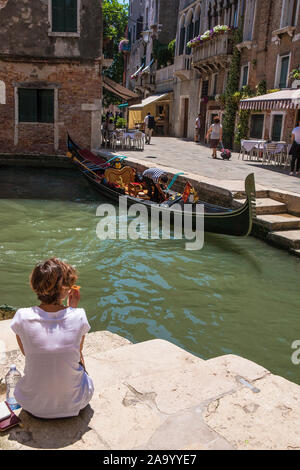 Ein Mädchen sitzt auf der Treppe von einer venezianischen Kanal mit einer Gondel gegenüber: Rio del Megio und Calle Larga, Santa Croce, Venedig, Italien Stockfoto