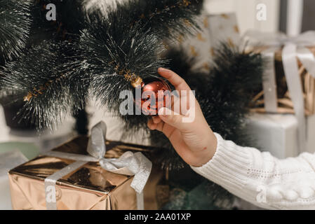 Eine Hand in einem weißen Strickpullover hängt ein Red christmas Ball auf dem Zweig eines künstlichen Weihnachtsbaum. Ein Schuß von Dekoration Weihnachten t Stockfoto