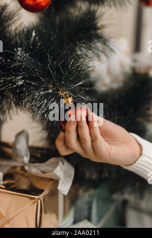 Eine Hand in einem weißen Strickpullover hängt ein Red christmas Ball auf dem Zweig eines künstlichen Weihnachtsbaum. Ein Schuß von Dekoration Weihnachten t Stockfoto