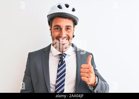 Junge gut aussehender Geschäftsmann Fahrradhelm und drahtlose Kopfhörer über isolierte Hintergrund tun glücklich Daumen nach oben Geste mit der Hand. Die Genehmigung der Ausdruck Stockfoto