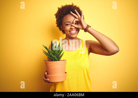 Junge afrikanische amerikanische Frau mit Cactus pot über isolierte gelber Hintergrund mit glücklichen Gesicht lächelnd tun ok Zeichen mit der Hand am Auge durch Suchen Stockfoto