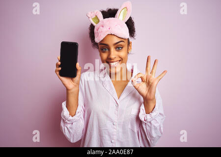 Afro Frau in Pyjama und Maske angezeigt Smartphone über isolierte rosa Hintergrund tun ok Zeichen mit den Fingern, ausgezeichnete Symbol Stockfoto