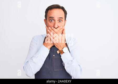 Mittleres Alter Ladenbesitzer Mann mit Schürze stehend über isoliert weißer Hintergrund schockiert über den Mund mit Händen für Fehler. Geheime Konzept. Stockfoto