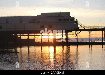 Aberystwyth, Wales, UK. Nov, 2019 18. Eine herrliche warme Winter tag auf der West Wales Küste, Sonnenuntergang bringt in einem murmuration Tausende von Staren in der Abenddämmerung von der Fütterung Felder um die walisische Küste auf der eisernen Beine der viktorianischen Badeort Pier zum roost. Credit: Mike Davies/Alamy leben Nachrichten Stockfoto