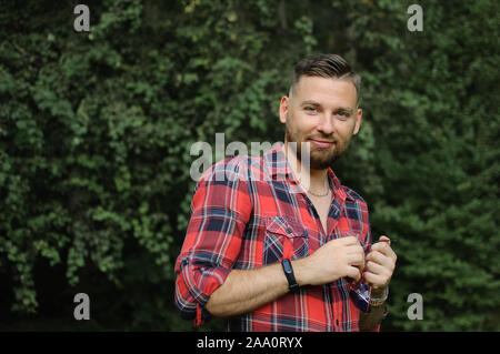 Brutale bärtiger junger Mann im roten T-Shirt steht im Freien auf grüne Bäume Hintergrund. Hüfthose style Konzept. Stockfoto