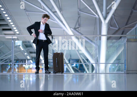 Junge Unternehmer stehen im Flughafen vor einem Glas guard Geländer mit seinem rollenden Fall etwas Suche in seinem Anzug Tasche Stockfoto