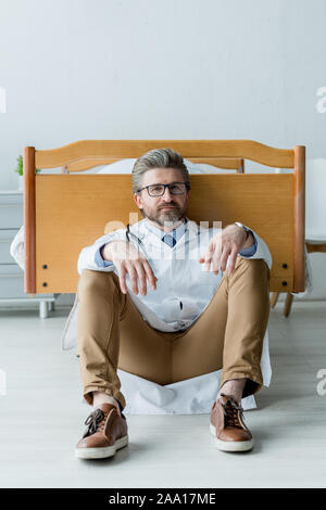 Stattliche Arzt im weißen Kittel sitzen auf dem Boden und Kamera im Krankenhaus Stockfoto