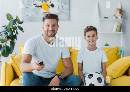 Freundliche Vater holding Remote Controller in der Nähe von Son mit Fußball sitzen auf einem Sofa Stockfoto