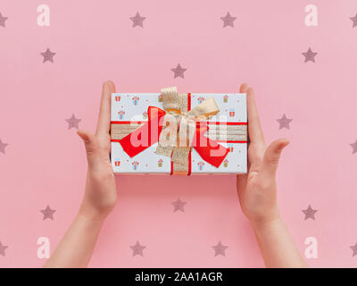 Geschenk Box Startseite anzeigen Foto Mädchen hält in den Händen einer kleinen Geschenkkarton mit einer Gold- und rote Schleifen auf einem Rosa dekorativen Hintergrund gebunden Stockfoto