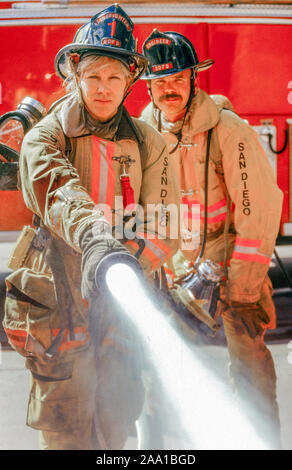 Ihre Sicherheit tragen, Mantel und Helm, eine Frau fire fighter zeigt ihre Fähigkeit mit einem Feuerwehrschlauch neben ihr Rig in der Gesellschaft von einem männlichen Kollegen Downtown San Diego, CA. Stockfoto