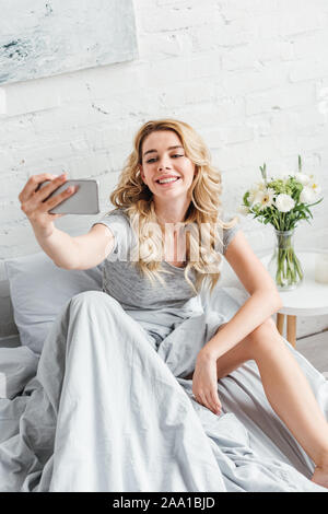 Selektiver Fokus der fröhliches Mädchen, die in der Nähe von selfie Vase mit Blumen auf dem Nachttisch Stockfoto