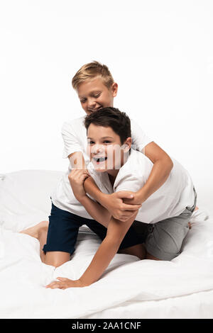 Fröhliche Junge lächelnd umarmen, Bruder, während Spaß auf dem Bett isoliert auf weißem Stockfoto