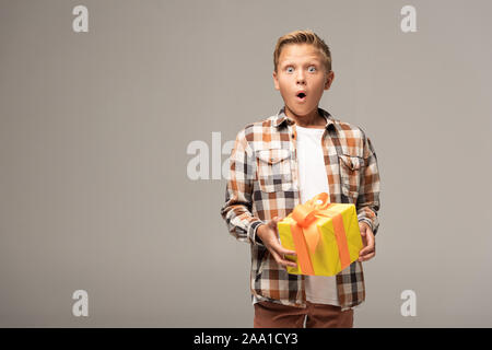 Schockiert boy Holding gelb Geschenkbox und mit Blick auf die Kamera isoliert auf Grau Stockfoto