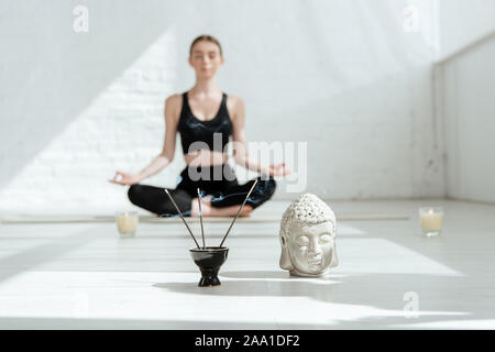 Selektiver Fokus der junge Frau sitzt in der halben Lotussitz in der Nähe von Buddha head Skulptur, aromatische Sticks und Kerzen Stockfoto