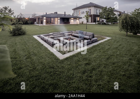 Tolle, moderne Immobilien mit einem zweistöckigen Haus, Spa Gebäude, grüne Wiese mit Bäumen und einem Relax Zone mit einer Feuerstelle auf dem Hintergrund der bewölkten Himmel. Stockfoto