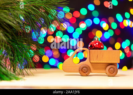 Eine Nahaufnahme eines Holz- Lkw brachte roten Weihnachtskugeln zu einem Weihnachtsbaum auf dem Hintergrund der vielen bunten Lichtern. Bild für das Neue Jahr Design, Stockfoto