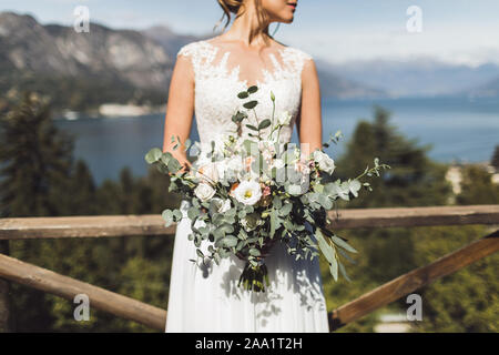 Woman Holding in den Händen große Hochzeit Blumenstrauß im rustikalen Stil. Grünen und weißen Rosen. Blick auf den See und die Berge im Hintergrund. Stockfoto