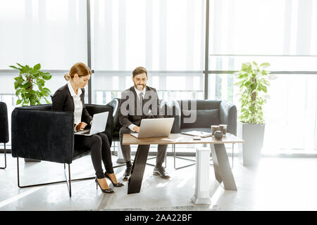 Angestellte von Finanzinstituten arbeiten mit Laptops in der luxuriösen Büro, sitzen auf dem bequemen Sofa in der Nähe der grossen Fenster Stockfoto