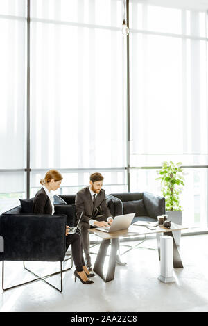 Angestellte von Finanzinstituten arbeiten mit Laptops in der luxuriösen Büro, sitzen auf dem bequemen Sofa in der Nähe der grossen Fenster Stockfoto