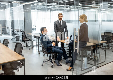 Gruppe von streng gekleidete Geschäftsleute in einer Diskussion, sitzen am Arbeitsplatz mit einem Computer im modernen Büro mit Glas Partitionen Stockfoto