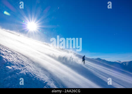 Steile Skipiste in windigen, sonnigen Wetter. Ein einsamer unkenntlich Snowboarder steigt herab und nimmt eine Menge Schnee Staub im Gegenlicht der Sonne Stockfoto