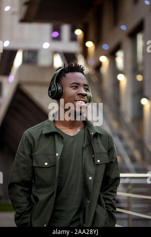 Lächelnd afrikanisch-amerikanische Mann im eleganten Jacke in drahtlosen Kopfhörern Musik hören auf der Straße von den Abend Stadt Stockfoto