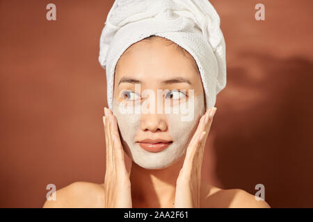 Portrait von jungen asiatischen Frau auf orangefarbenem Hintergrund mit Lehm Maske im Gesicht und ein Handtuch auf dem Kopf lächelnd. Stockfoto