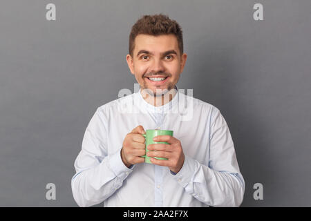 Freestyle. Junger Mann Studio stehen isoliert auf Grau trinken heißen Kaffee Lächeln glücklich Stockfoto