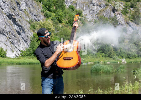 Foto von schreienden Stoppeln fröhliche Schöne attraktive Musiker gegen die Kulisse der hellen Sommer Landschaft. junge Hipster bärtigen Kerl spielt den Git Stockfoto