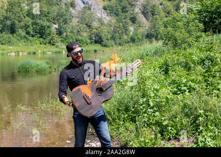 Foto von schreienden glücklich aussehenden Musiker gegen die Kulisse der hellen Sommer Landschaft. junge Hipster bärtigen Kerl brennende Gitarre hält. Stockfoto