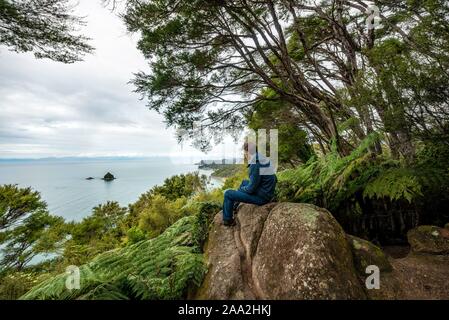 Junger Mann sitzt auf Felsen und blickt auf das Meer, Abel Tasman National Park, South Island, Neuseeland Stockfoto