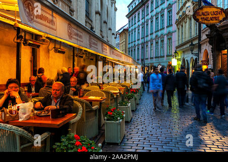 Menschen in beleuchteten Outdoor Restaurant sitzt neben Menge Straße mit Kopfsteinpflaster in der Altstadt von Prag. Stockfoto