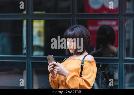 Schöne junge Mädchen in modische Kleidung verwendet Smartphone mit schwarzem Hintergrund Windows im Stehen. Stockfoto