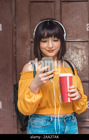 Schöne junge Mädchen in modische Kleidung Musik hören mit Kaffeetasse, während standind Stockfoto