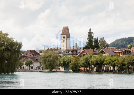 Unterseen, Interlaken, Schweiz: Bäume säumen das Ufer der Aare, hinter dem sich Turm und Uhr der alten Kirche über den Dächern erheben. Stockfoto