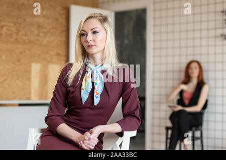 Charmante blonde Frau in rotem Kleid sitzt im Stuhl. Eine andere Frau sitzt im Stuhl auf Hintergrund. Selektive konzentrieren. Stockfoto
