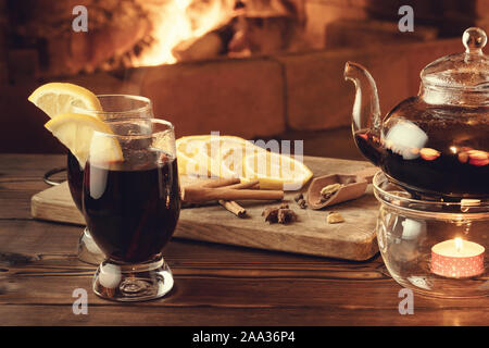 Zwei Gläser von Glühwein und Teekanne auf einem holztisch vor einem brennenden Kamin. Stockfoto