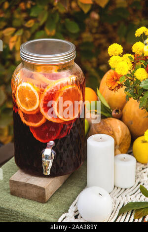 Glühwein in einem Glaszylinder auf einem Tisch im Garten für ein festliches Abendessen mit der Familie neben Kerzen, Kürbisse und Blumen. Einrichtung und Tabelle einstellen. Stockfoto