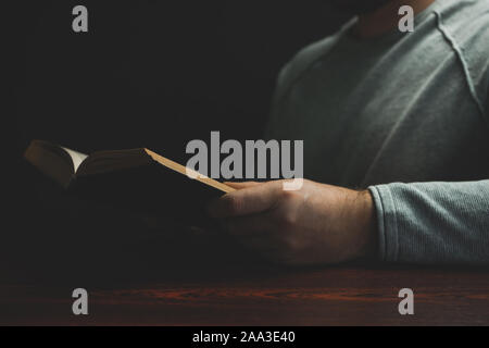 Junger Mann saß auf einem roten hölzernen Tisch lesen Das alte Buch und schwarzem Hintergrund. Stockfoto