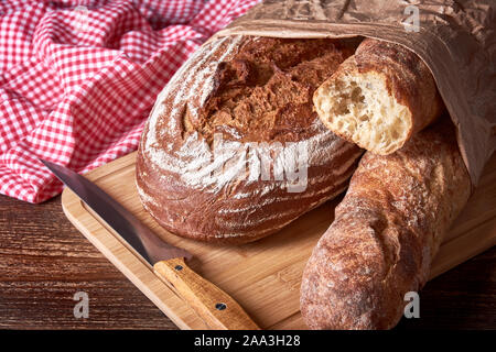 Laib Brot, Baguette und Gebäck auf hölzernen Tischplatte mit scharfen Küchenmesser. Rotes Tuch im Hintergrund. Stockfoto