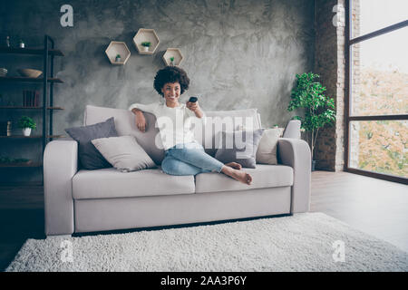 Foto ziemlich dunkle Haut wellig Lady heimelige Stimmung halten TV-Fernbedienung ändern Kanal Suche Favoriten Humor zeigen Sitzen gemütliche Couch lässig Stockfoto