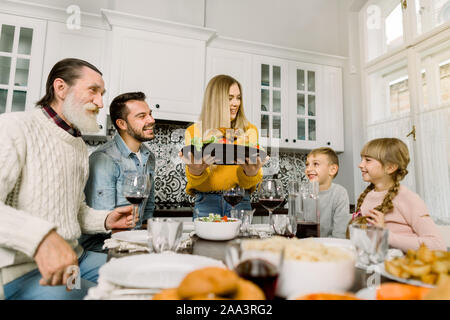 Die Familie setzt sich zum Abendessen auf Thanksgiving. Junge Frau dient eine festliche Türkei mit Salat, Großvater, Vater und Kinder sitzen und schauen sie sich Stockfoto