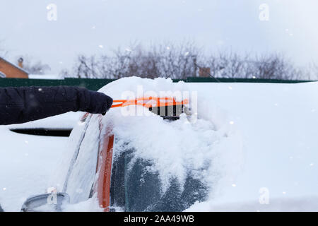 Bürste in mans hand. Man löscht Windschutzscheibe von orange Auto vom Schnee. Auto mit Schnee bedeckt. Winter Probleme der Autofahrer. Snowy. Stockfoto