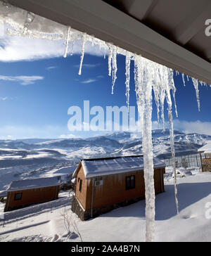 Dach mit großen sonnendurchfluteten Eiszapfen und kleinen Holzhäuser an verschneite Berge. Kaukasus, Shahdagh, Aserbaidschan. Weitwinkelaufnahme. Stockfoto