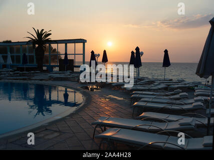 Liegestühle und Sonnenschirme am Strand mit Blick auf den randlosen Pool und Strand bei Sonnenuntergang in einem Resort. Die Sonne im Meer. Die Insel Rhodos. Dodekanes Griechenland Stockfoto