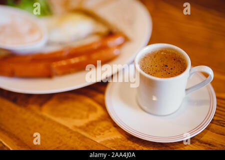 Klassische englische Frühstück mit Espresso in weiße Krug, zwei Würstchen, Rührei, Toast, Salat auf Holz- Hintergrund. Stockfoto