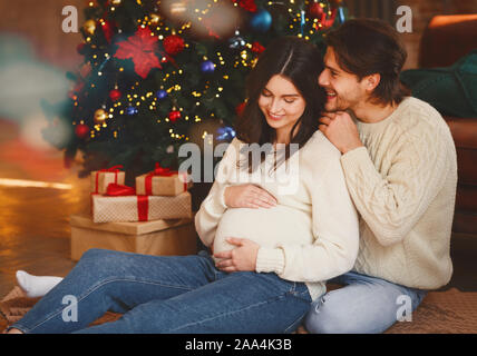 Die schwangere Frau und ihr Mann sitzen durch Weihnachtsbaum Stockfoto