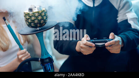 Mann hält Gamepad in der Hand, im Hintergrund Rauch von Wasserpfeifen. Stockfoto