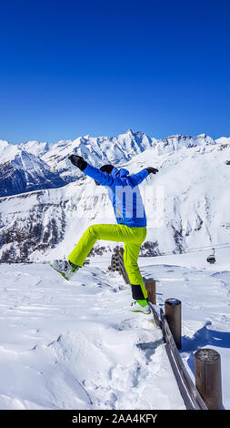 Mann in einem Skifahren Outfits in den frischen Schnee springen. Der Mensch trägt blaue Jacke und neon grün Hose. Viel frischem Pulverschnee um ihn herum. Hohe Mou Stockfoto